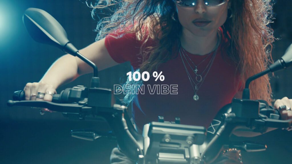 Nahaufnahme von Eli Preiss am Motorradlenker mit Sonnenbrille und rotem Shirt unter dem Claim 