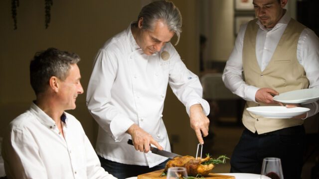 Unsere Restaurant Branding Agentur dokumentiert wie Chef Richard Brunnauer den Braten am Tisch tranchiert und dabei die persönliche Gastfreundschaft seines Drei Hauben Restaurants zeigt. Chef Richard Brunnauer tranchiert einen Braten am Gästetisch im Restaurant Brunnauer als Teil unseres Foto und Branding Projekts für Gastronomie von Wuger Hospitality