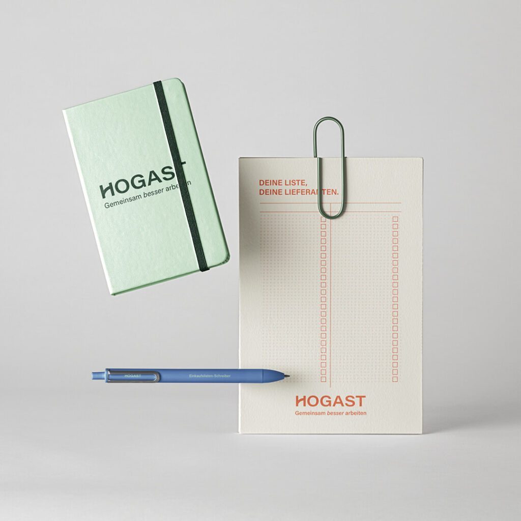 „Gestaltete HOGAST Give-aways bestehend aus grünem Notizbuch, Kugelschreiber und Lieferantenliste mit neuem Logo und Claim – Design- und Brandingprojekt von WUGER Hospitality im Rahmen des HOGAST Rebrandings.