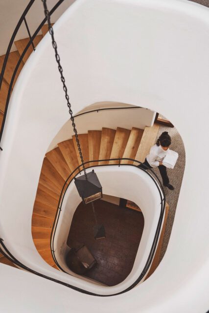 Blick von oben auf eine elegante Wendeltreppe mit Holzstufen und weißem Geländer, eine Hotelmitarbeiterin trägt Handtücher – Interior-Fotoproduktion für das HOGAST Rebranding Projekt von WUGER Hospitality im Bereich Hotellerie und Markenauftritt
