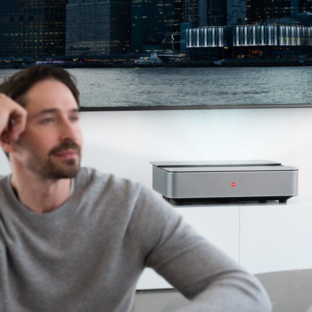 Ein Mann sitzt vor dem Leica Cine 1 Projektor, der ein urbanes Nachtpanorama an die Wand projiziert – modernes Home-Cinema-Erlebnis von WUGER.