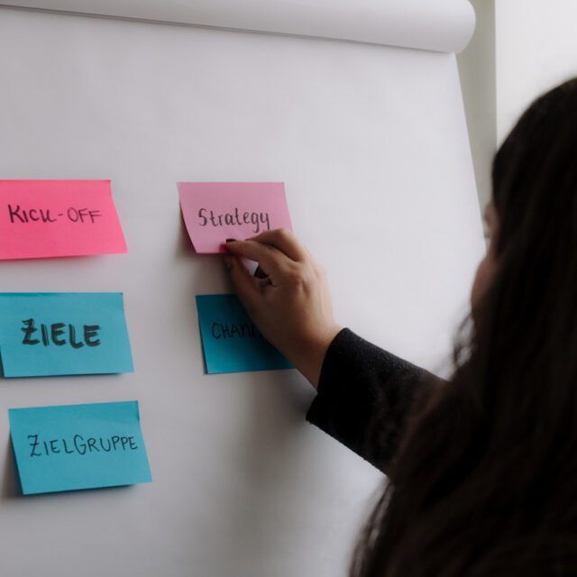 Wir denken Marken strategisch, um den Emotionen in ihr Richtung zu geben. Mitarbeiterin klebt im Strategie Meeting zur Markenanalyse und Digitalstrategie Post-Its an Board