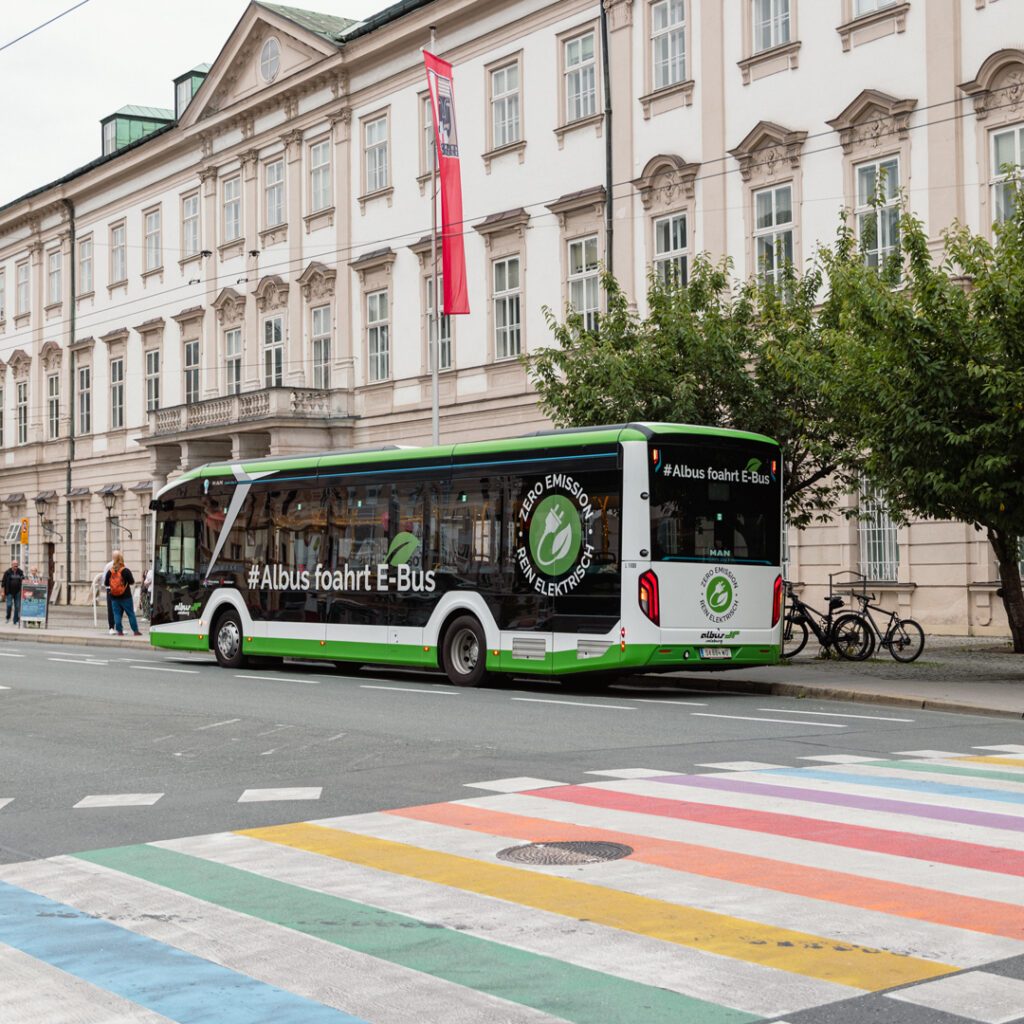 Ein Ort, an dem Vielfalt mitfährt und Begegnungen selbstverständlich Raum bekommen. Ein Elektrobus hält an einem Zebrastreifen in Regenbogenfarben und setzt ein sichtbares Zeichen für Vielfalt, Offenheit und queere Begegnungen im öffentlichen Raum