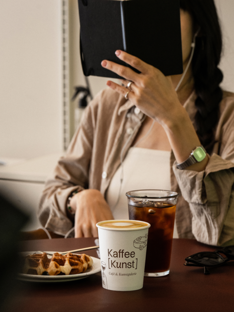 Wuger zeigt Branding auf To Go Bechern neben Gebäck auf Tisch mit Kunden für die Markenbildung von Kaffee Kunst
