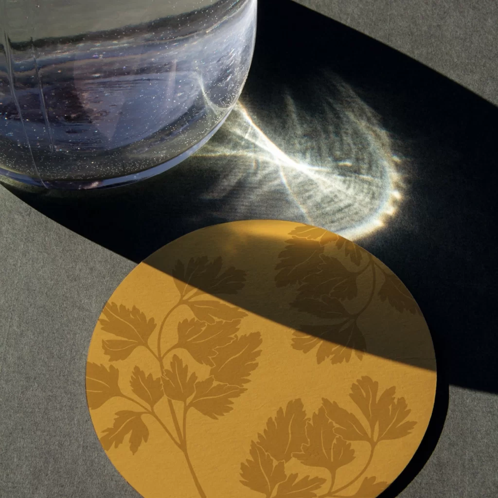 Sogar der Untersetzer erzählt die Markengeschichte mit, und genau solche Details bleiben den Gästen in Erinnerung. Goldener Untersetzer mit Kräuterillustration und Glas im Streiflicht als Gastronomie-Branding und Corporate Design von Wuger