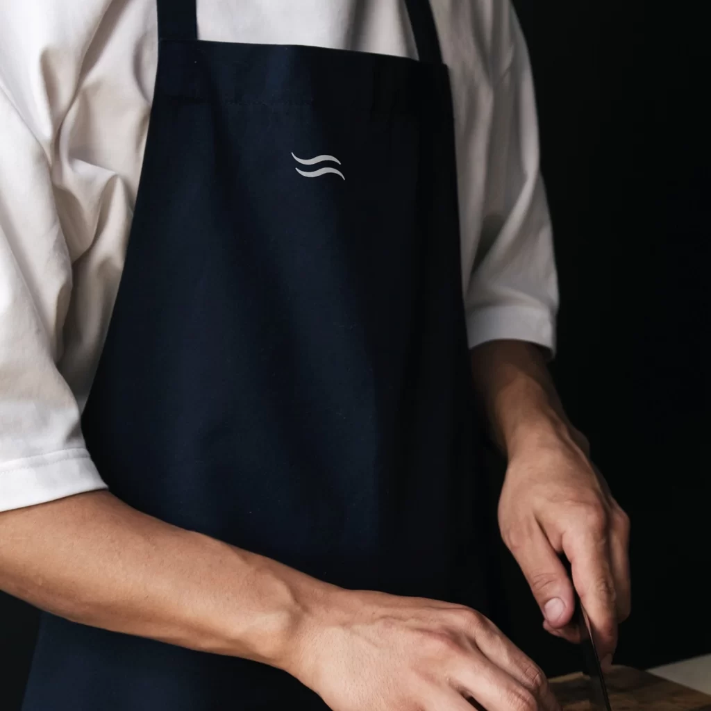 Selbst die Schürze in der Küche trägt das Zeichen der Marke, ganz beiläufig und trotzdem unverkennbar. Koch in dunkler Schürze mit gesticktem Wellenlogo als Corporate Design und Logo von Wuger, Messer griffbereit in der Hand